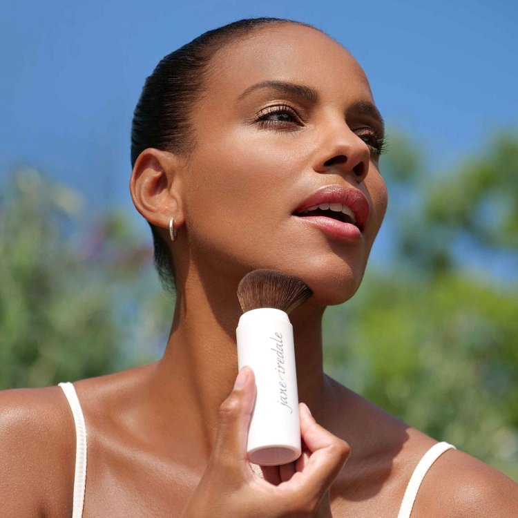 Na fotografii je vidět ženu, která nanáší pudr pomocí štětce Jane Iredale Powder-Me SPF. Produkt má formu praktického aplikátoru se štětcem a používá se venku za slunečného dne, což podtrhuje jeho funkci ochrany před slunečním zářením (SPF).