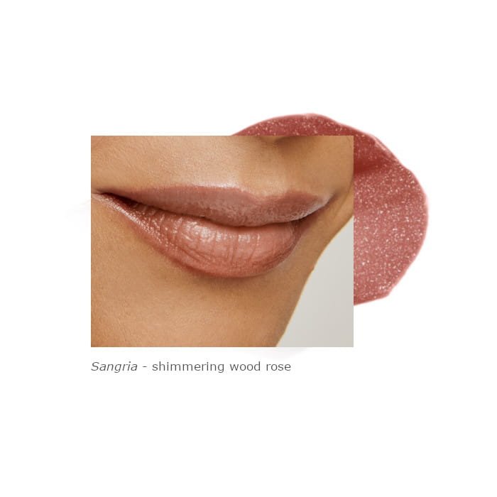 Na obrázku jsou rty modelky nalíčené odstínem Sangria. Produkt má teplý, tlumený růžovo-hnědý odstín s jemným leskem. Textura je lesklá s drobnými třpytkami, které dodávají rtům zářivý efekt.