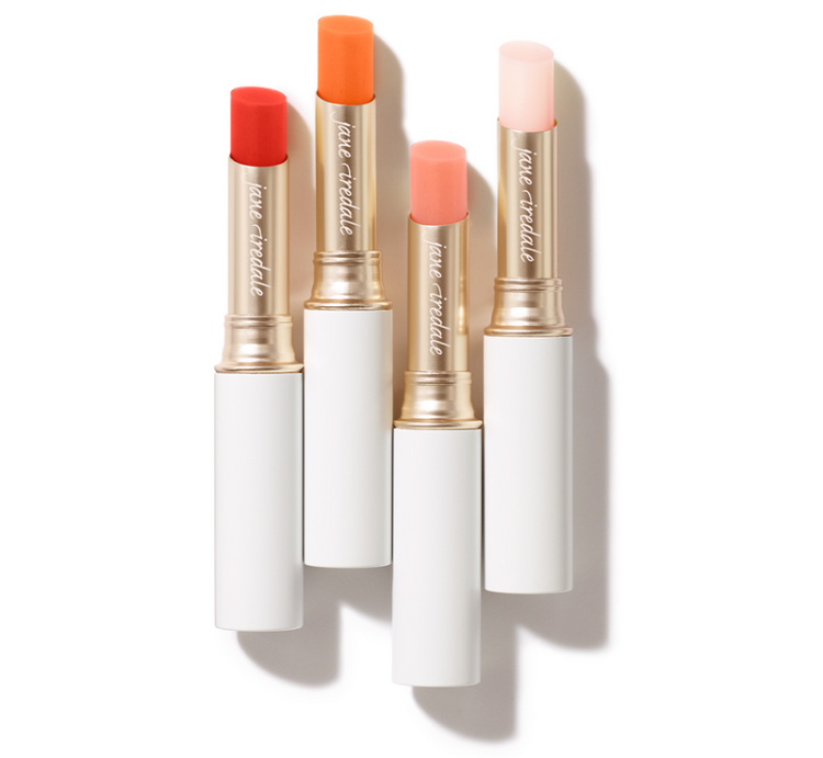 Na obrázku jsou vidět čtyři rtěnky z kolekce Just Kissed Lip and Cheek Stain značky Jane Iredale. Produkty jsou v elegantních bílých obalech se zlatým zakončením. Každá rtěnka má jiný odstín.