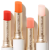 Na obrázku jsou čtyři ochranné rtěnky značky Jane Iredale ve formě tyčinky. Každá má jiný odstín: intenzivně červený, energický oranžový, jemný broskvový a transparentní růžový. Obaly jsou elegantní, zlatobílé a lesklé.