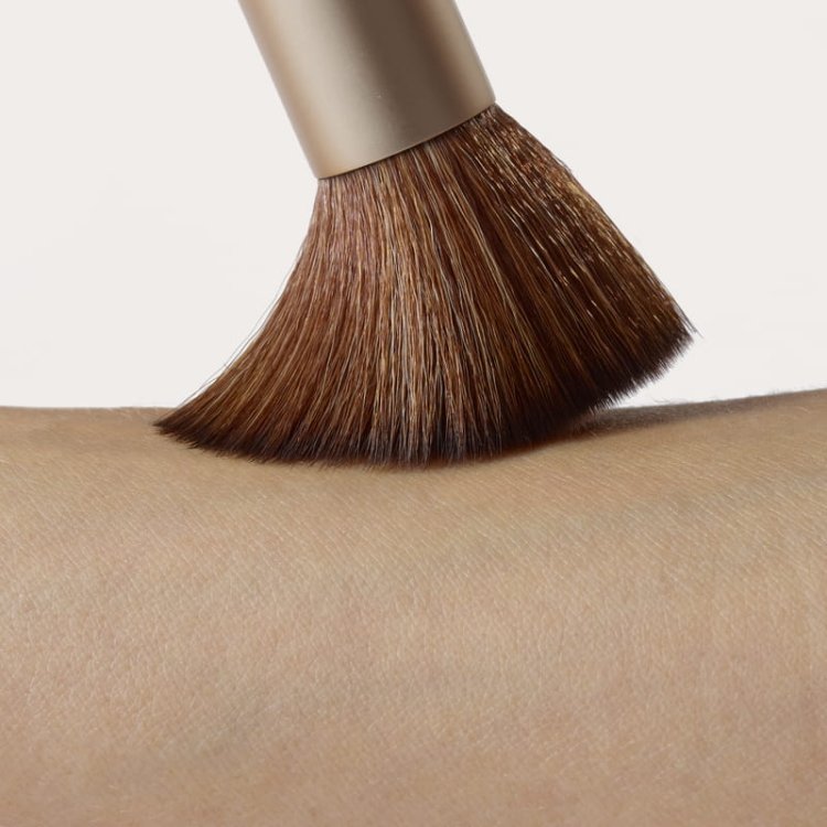 Na obrázku je vidět štětec na make-up Handi jemně klouzající po pokožce. Štětiny jsou husté, měkké a tvarované, s tmavší základnou a světlejšími konečky.