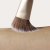 Na obrázku je detail štětce na make-up přejíždějícího po pokožce. Štětiny jsou měkké, husté, se světlými konečky a hnědou základnou, a objímka má saténový zlatý povrch. Štětec se jemně dotýká pokožky, čímž zdůrazňuje svou nadýchanost a pružnost.