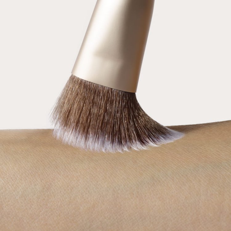 Na obrázku je detail štětce na make-up přejíždějícího po pokožce. Štětiny jsou měkké, husté, se světlými konečky a hnědou základnou, a objímka má saténový zlatý povrch. Štětec se jemně dotýká pokožky, čímž zdůrazňuje svou nadýchanost a pružnost.