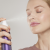 Na fotografii žena si stříká na obličej hydratační mlhu Lavender Hydration Spray od Jane Iredale. Má zavřené oči a uvolněný výraz ve tváři. Produkt, který drží v ruce, je červená lahvička s bílým rozprašovačem – ideální pro citlivou pleť.