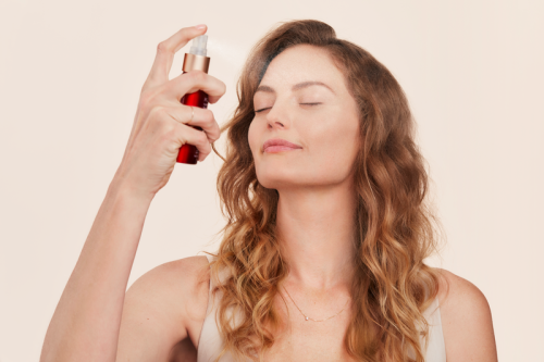 Na fotografii žena si stříká na obličej hydratační mlhu Pommist Hydration Spray od Jane Iredale. Má zavřené oči a uvolněný výraz ve tváři. Produkt, který drží v ruce, je červená lahvička s bílým rozprašovačem – ideální pro všechny typy pleti.