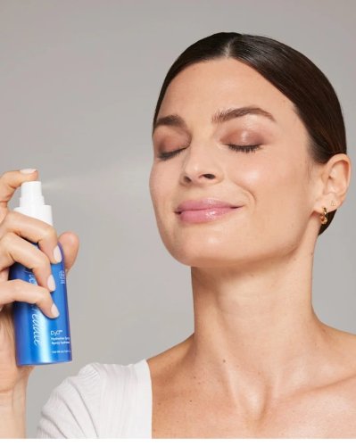 Na fotografii žena si stříká na obličej hydratační mlhu D₂O™ Hydration Spray od Jane Iredale. Má zavřené oči a uvolněný výraz ve tváři. Produkt, který drží v ruce, je modrá lahvička s bílým rozprašovačem – ideální pro suchou pleť.