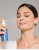 Na fotografii žena si stříká na obličej hydratační mlhu D₂O™ Hydration Spray od Jane Iredale. Má zavřené oči a uvolněný výraz ve tváři. Produkt, který drží v ruce, je modrá lahvička s bílým rozprašovačem – ideální pro suchou pleť.
