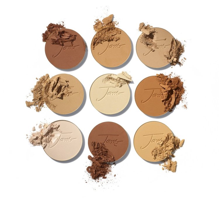 Na obrázku je vidět devět kulatých lisovaných pudrů Jane Iredale v různých odstínech – od velmi světlých po tmavé, ideálních pro přizpůsobení různým tónům pleti. Některé z nich jsou lehce rozdrcené, což zdůrazňuje texturu produktu.