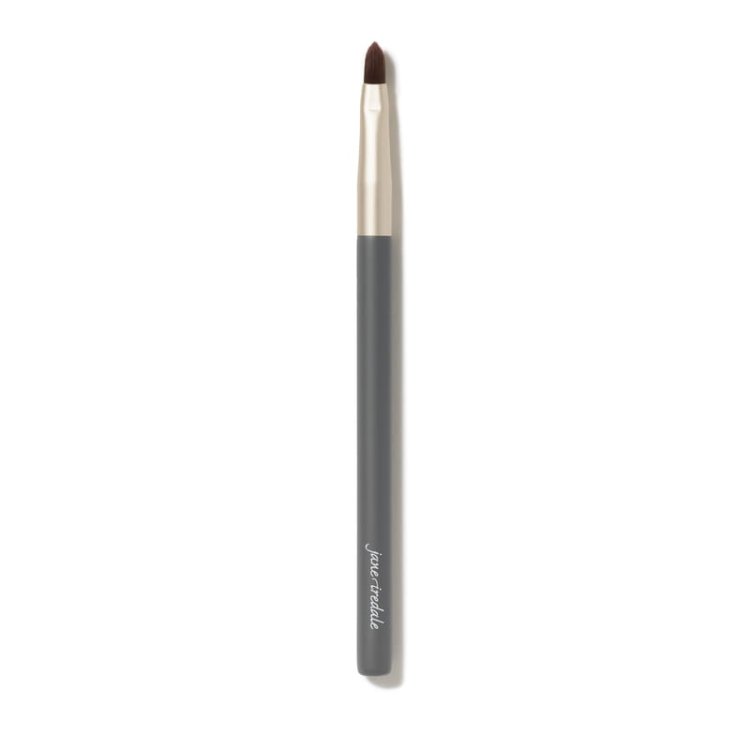 Na obrázku je tenký štěteček na make-up značky jane iredale. Má štíhlý tvar, šedou rukojeť s logem značky a zlatou objímku. Štětiny jsou krátké, zastřižené do špičky, což z něj dělá ideální nástroj pro precizní aplikaci produktů.