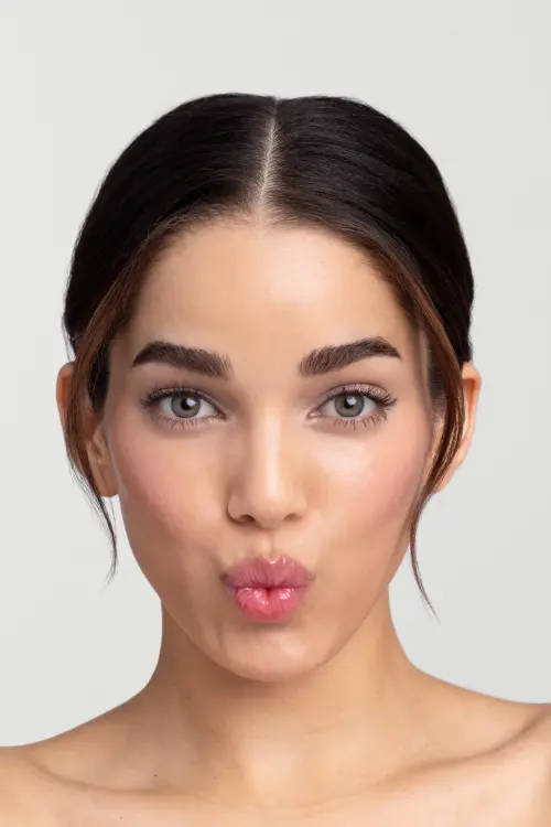 Fotografie zachycuje detail tváře modelky s tmavými, sepnutými vlasy a zelenýma očima. Modelka se dívá přímo do objektivu a špulí rty. Make-up je jemný, se zvýrazněnými řasami, rozjasněnou pletí a růžovými, lesklými rty.