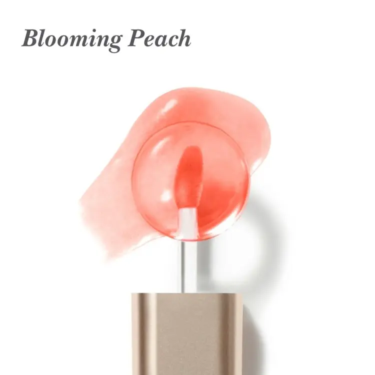 Na bílém pozadí je aplikátor oleje na rty Jane Iredale Just Kissed Lip Oil s měkkou, zaoblenou špičkou v korálovém odstínu. V pozadí je vidět rozetřený vzorek oleje v broskvově korálové barvě s transparentní, lesklou texturou.