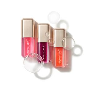 Na bílém pozadí jsou tři oleje na rty Jane Iredale Just Kissed Lip Oil v průhledných obalech s metalickými zlatými uzávěry. Oleje mají odstíny intenzivní růžové, bobulově fialové a korálové.