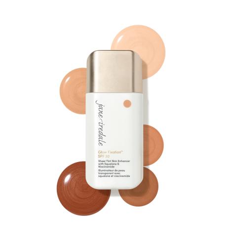 Na obrázku je Jane Iredale Glow Fixation SPF 40 Sheer Tint Skin Enhancer v bílém balení s aplikátorem. Kolem produktu jsou zobrazeny vzorky kosmetického přípravku v několika tónech – od světlých béžovo-broskvových až po hlubší, teplé hnědé odstíny.