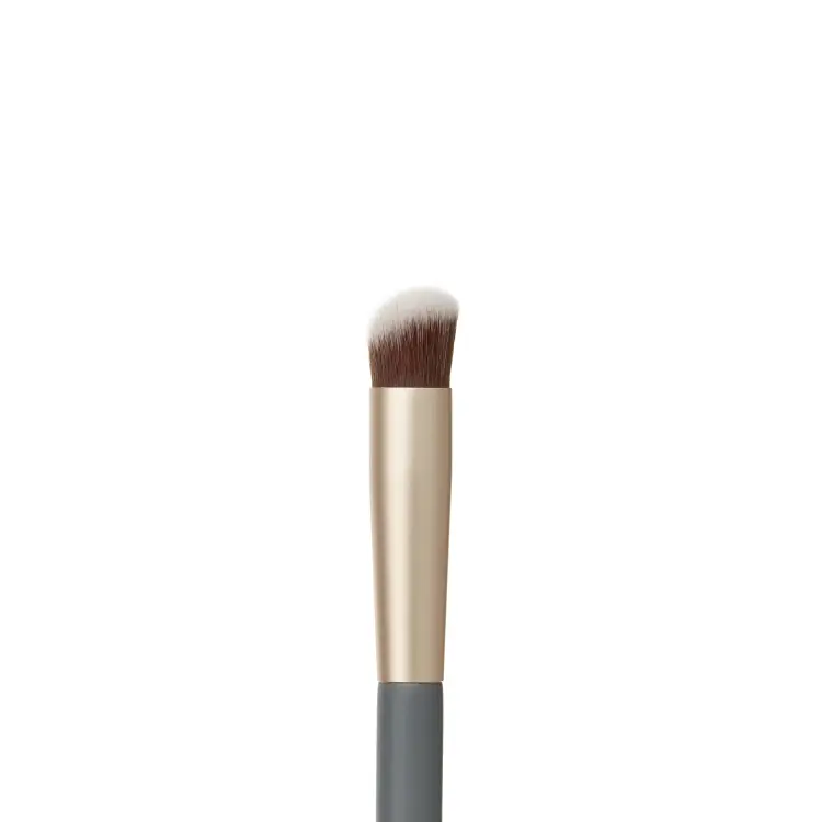 Fotografie zobrazuje detail hlavice štětce na korektor Jane Iredale Concealer Brush na bílém pozadí. Viditelná jsou hustá, mírně zkosená hnědá vlákna s bílými konečky, zasazená do zlaté objímky, která navazuje na šedou rukojeť.