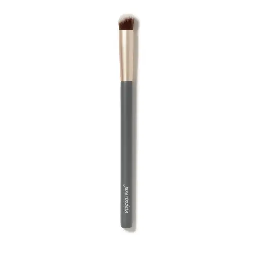 Fotografie zobrazuje štětec na korektor Jane Iredale na bílém pozadí. Má dlouhou šedou rukojeť s bílým logem značky, zlatou objímku a krátká, hustá, mírně zaoblená hnědá vlákna s bílými konečky.