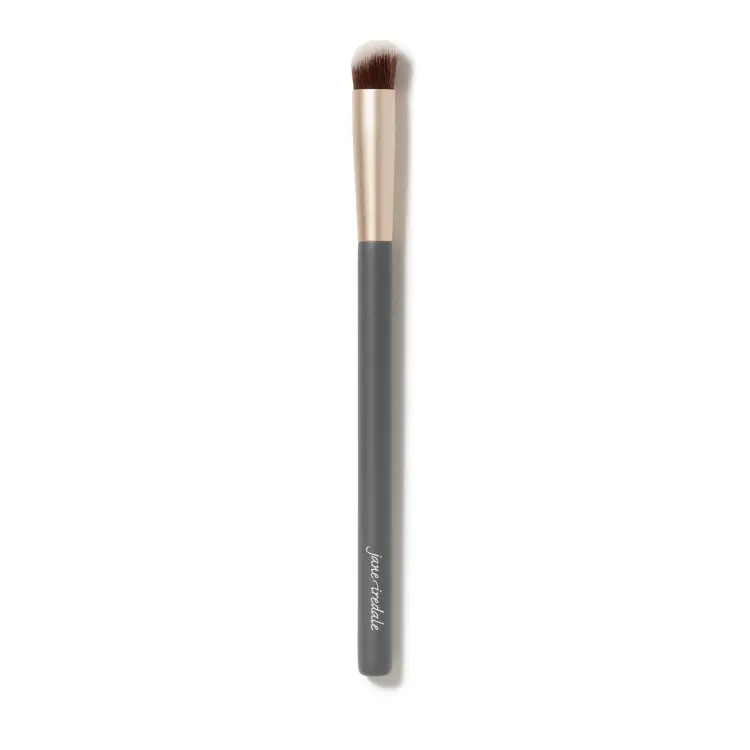 Fotografie zobrazuje štětec na korektor Jane Iredale na bílém pozadí. Má dlouhou šedou rukojeť s bílým logem značky, zlatou objímku a krátká, hustá, mírně zaoblená hnědá vlákna s bílými konečky.