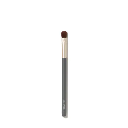 Fotografie zobrazuje štětec na líčení očí Jane Iredale Flat Eye Brush na bílém pozadí. Má dlouhou šedou rukojeť s bílým logem značky, zlatou objímku a krátká, hustá, plochá hnědá vlákna s mírně zaobleným zakončením.