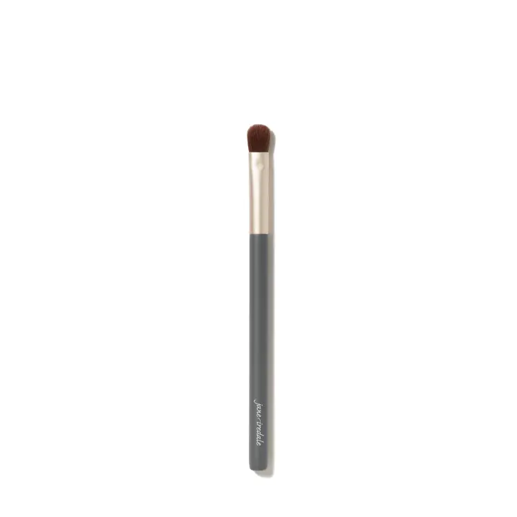 Fotografie zobrazuje štětec na líčení očí Jane Iredale Flat Eye Brush na bílém pozadí. Má dlouhou šedou rukojeť s bílým logem značky, zlatou objímku a krátká, hustá, plochá hnědá vlákna s mírně zaobleným zakončením.