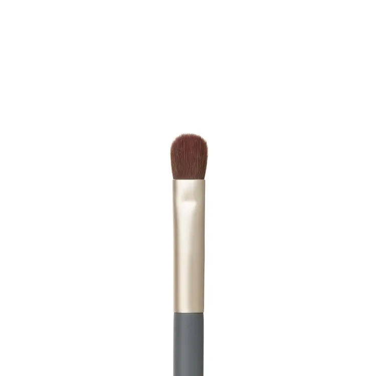 Fotografie zobrazuje detail hlavice štětce na líčení očí Jane Iredale Flat Eye Brush na bílém pozadí. Viditelná jsou krátká, hustá, plochá hnědá vlákna se zaobleným zakončením, zasazená do zlaté objímky, která navazuje na šedou rukojeť.