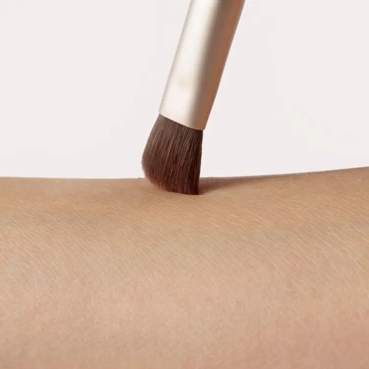 Fotografie zobrazuje detail štětce Jane Iredale Flat Eye Brush při kontaktu s pokožkou. Hustá, plochá hnědá vlákna se jemně dotýkají předloktí a ukazují tvar a preciznost hlavice štětce. Viditelná je také zlatá objímka.