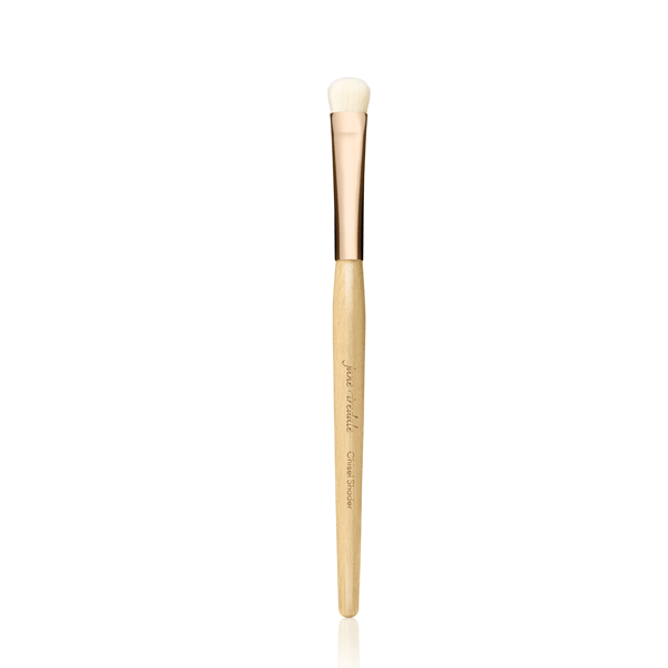 Na obrázku je vidět štětec na make-up značky Jane Iredale. Jedná se o tenký, precizní štětec s dlouhou dřevěnou rukojetí v přírodní barvě a elegantní zlatou objímkou. Hlava štětce je malá, zaoblená a vyrobená ze světlých štětin.
